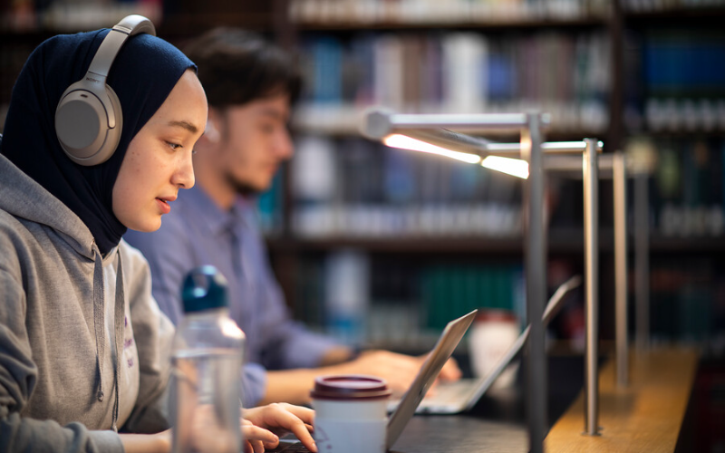 A UCL student wearing grey sweater, headphones and hijab in focus. Another student and water bottle not in focus.