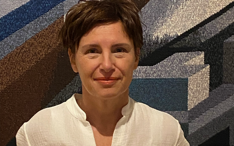 A woman with short brown hair, wearing a white shirt in front of a printed background
