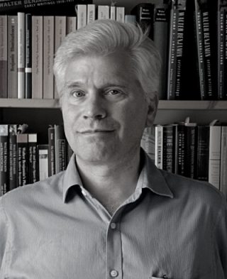 a man with light hair wearing a shirt stands in front of a bookcase