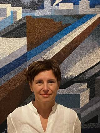 A woman with short brown hair, wearing a white shirt in front of a printed background