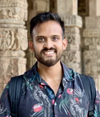 A young Indian man in a floral shirt smiles at the camera