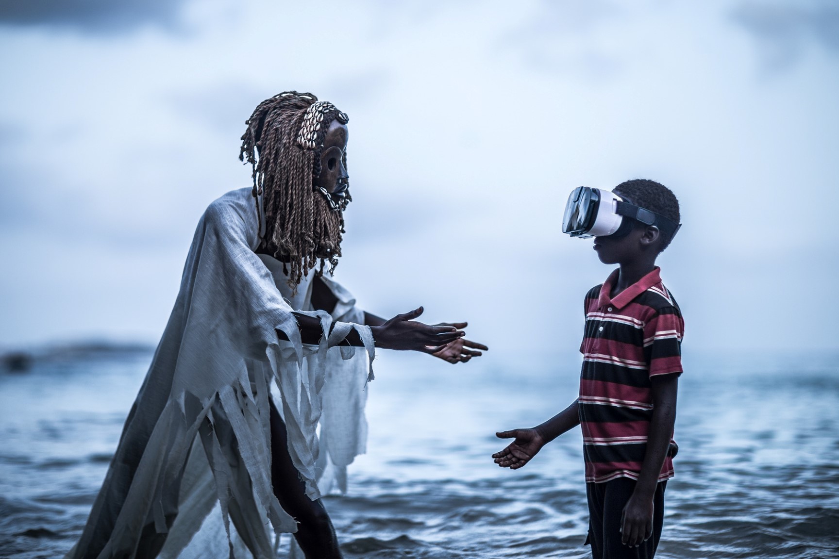 Pictured in this photographic artwork are two Black African figures facing one another, standing in front of a steely blue ripples of water that merges with a nebulous background at a vaguely distinguishable horizon.