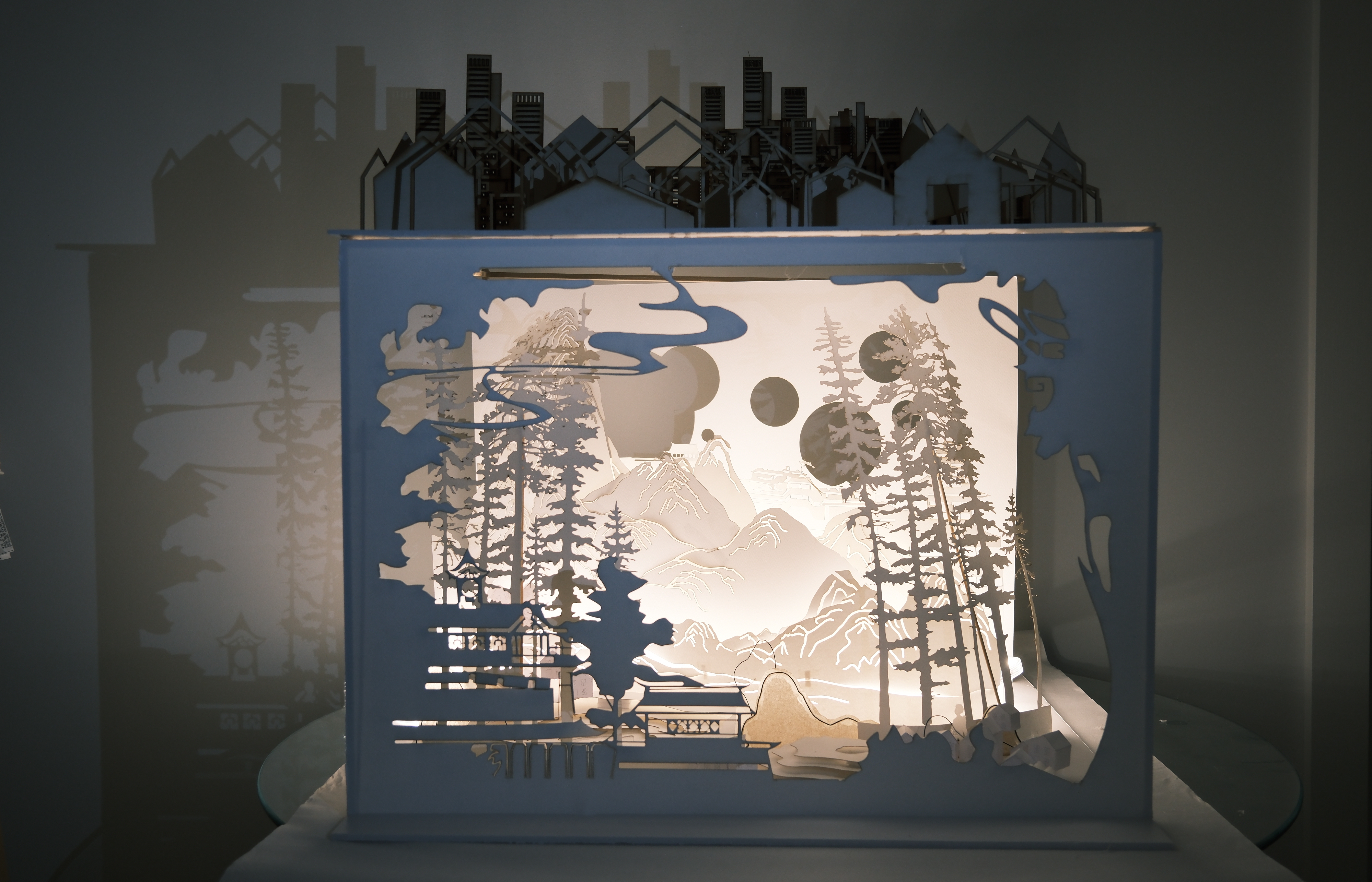 A paper cut out scene of landscape and city scape with shadow play on wall behind