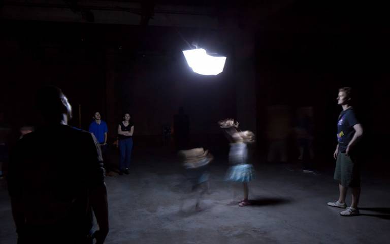 A bright light on the ceiling in a pitch black room with people below blurred through movement