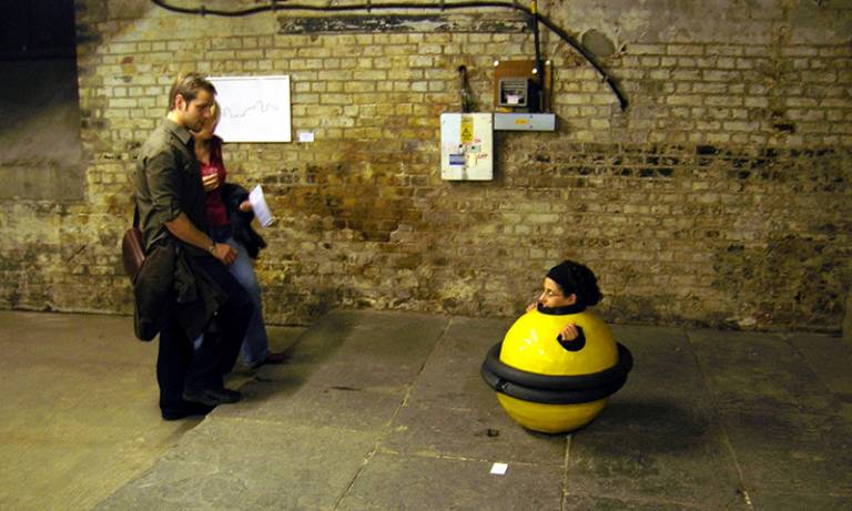 The image shows someone enclosed in a 'weeble' suit - a bright yellow human-sized plastic bubble - being looked at quizzically by two onlookers. It is a photograph of a performance by artist Noemi Lakmaier entitled 'Exercise in Losing Control'.
