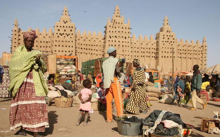 A colourful market place in Africa with women and children stood in front of a large sand building structure.