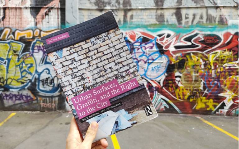 Hand holding a copy of the book Urban Surfaces against a graffitied wall