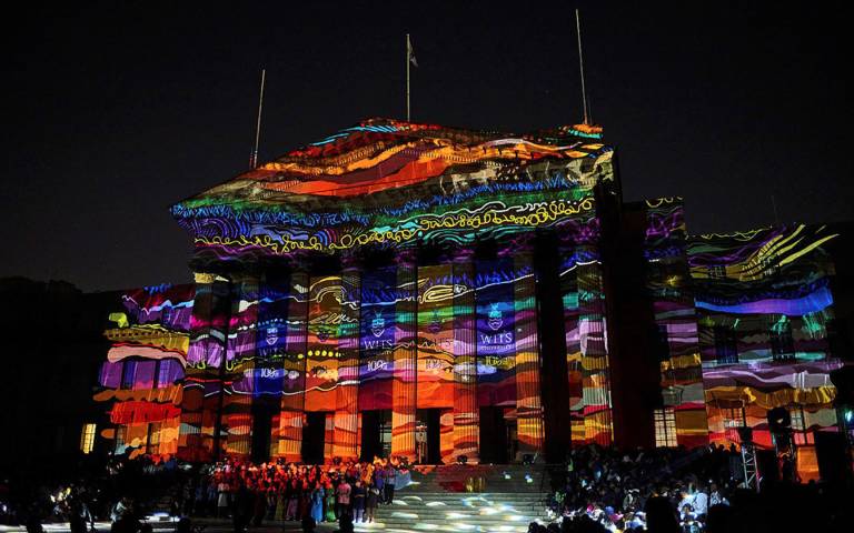 A photo of the Wits Great Hall during the Homecoming light show in 2022 by Brett Eloff. The building is illuminated with a colourful light graphic