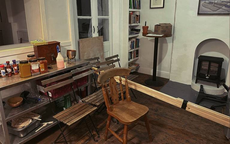A collection of wooden chairs and bound planks of wood arranged in a kitchen space in front of a fireplace with a wood stove and a metal food prep table with various food jars.