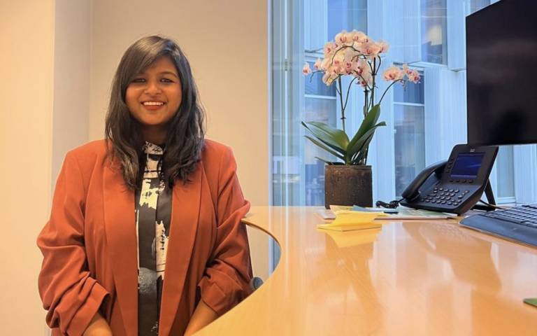 Ritwika Deb (UCL Alumni) smiling whilst standing next to a desk which has flowers a phone and a desktop on it.