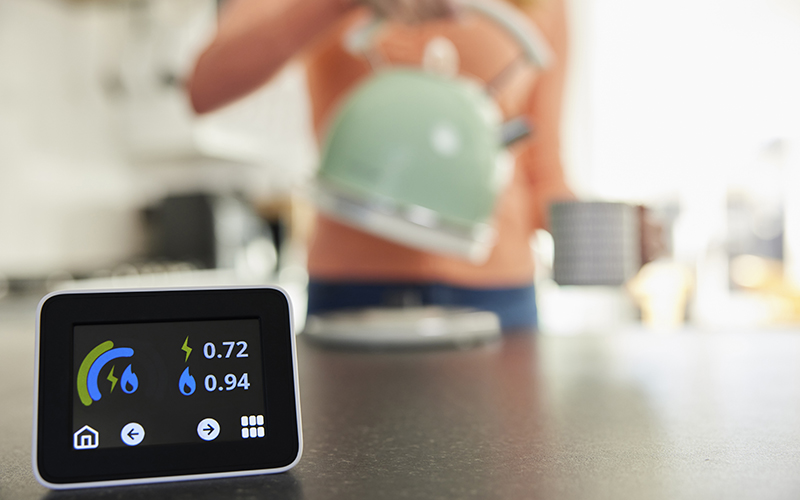 Photo of a smart meter with a person using a kettle in the background