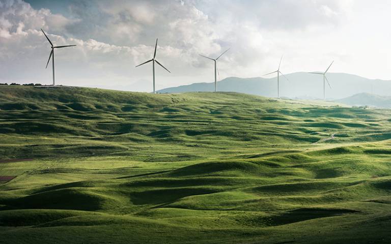 Photo shows a green hilly field with wind turbines in the background against a cloudy sky.