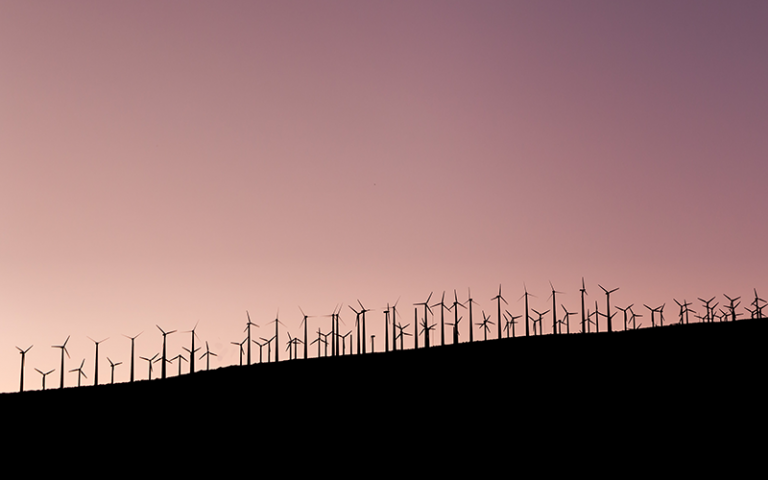 Photo shows silhouette of wind turbines against a pink sunset