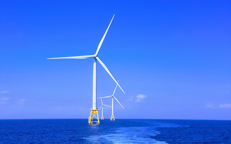 Photo shows a row of wind turbines stationed at sea with a blue sky behind