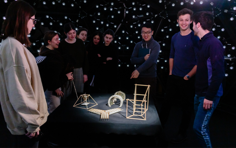 students stood in a dome covered in LED lights with one beam of light shining on small wooden models in the centre as they stand around in a semi-circle 