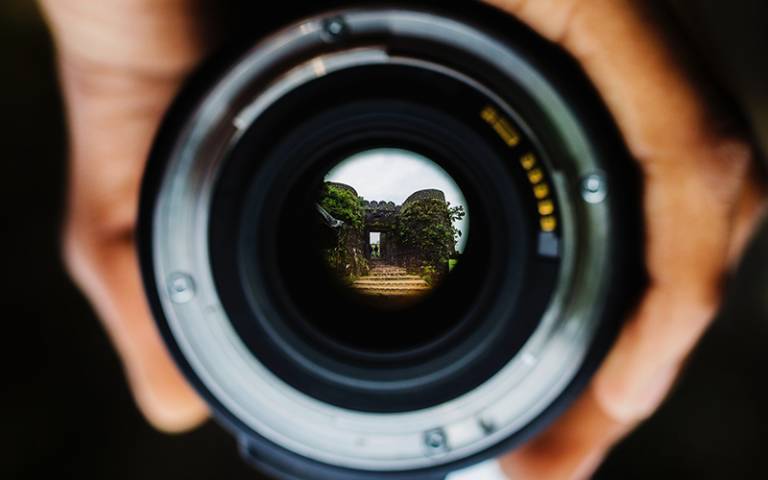 Photo shows hands holding a camera lens, the lens reflects a view of a castle