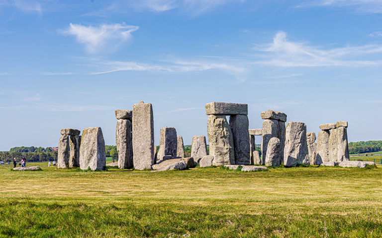 Photo shows Stonehenge site, a gray rock formation on a green grass field during daytime