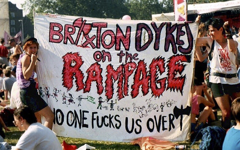 Two women hold up banner reading: Brixton dykes on the rampage, no one fucks us over. Park with protestors sitting.