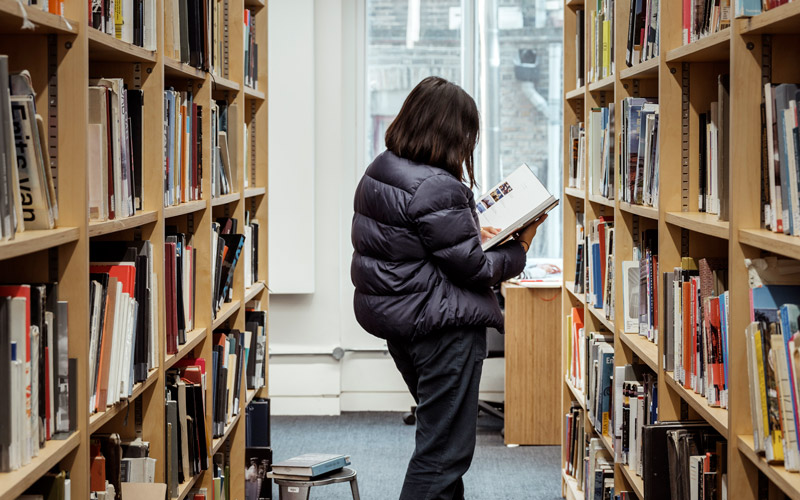 A person studies a book while standing between two shelves of library books