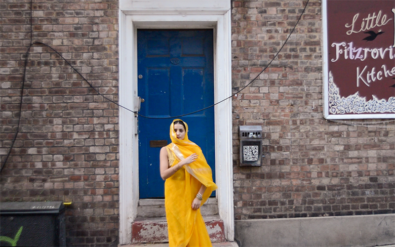 Woman in Indian dress standing in front of blue door, with sign reading: Little Fitzrovia Kitchen