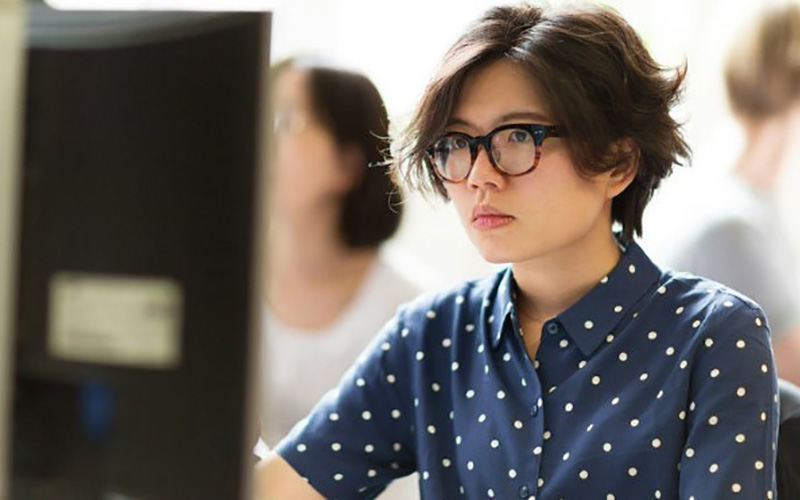 Woman with short dark brown hair and black thick rimmed glasses working on desktop computer wearing a button down polka dot short sleeved shirt