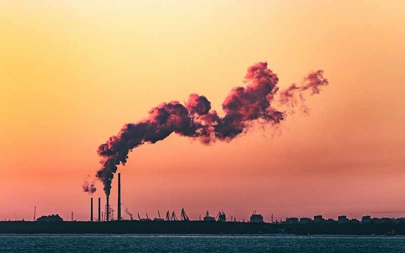 body of water under cloudy sky during sunset showing a factory in the distance polluting fossil fuels into the air in a big plume of smoke