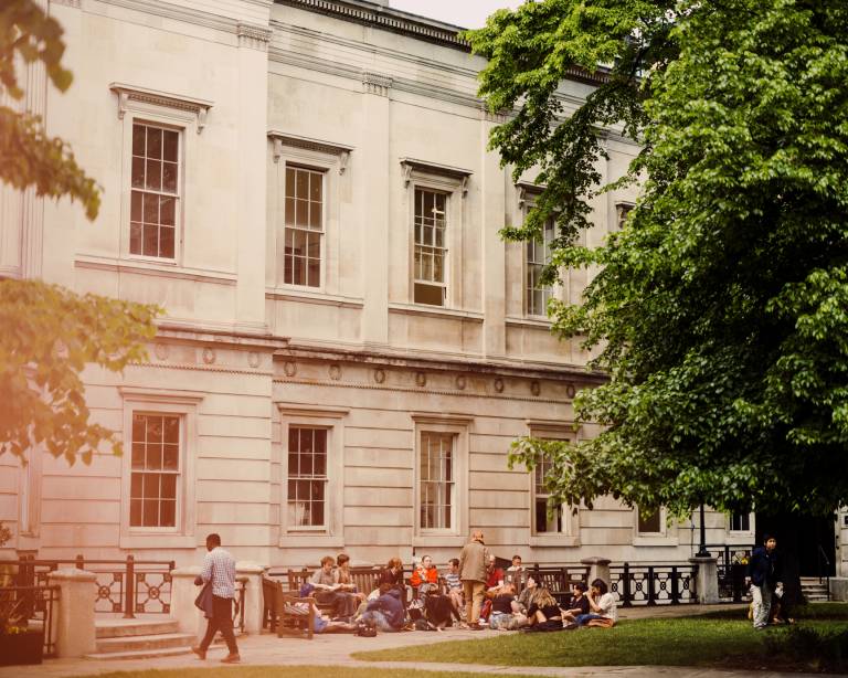 Image of UCL Bloomsbury campus showing a group of people walking into the building, sitting on benches and the ground