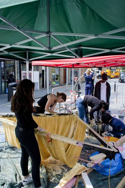Group of five students outside under tent, constructing wooden triangular frame with clamps and yellow fabric draped inside frame. 