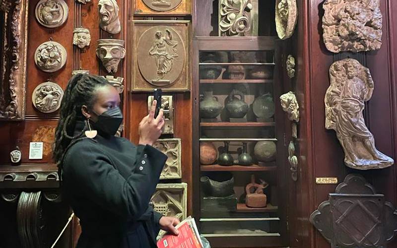 woman in facemask in museum of antiquities (pots and masks) takes photo with phone