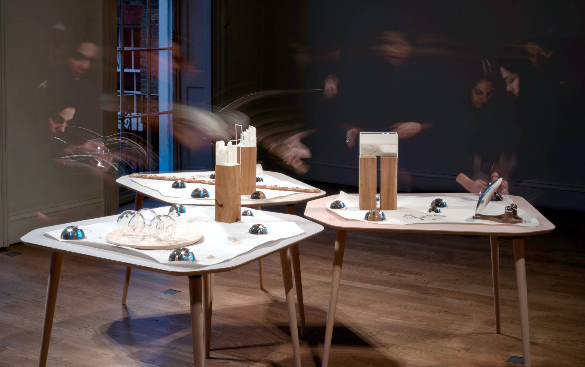 Three tables facing each other, with various pieces of white paper, glass, metal and wood figures. Long exposure shows a women's figure placing the items on the table.