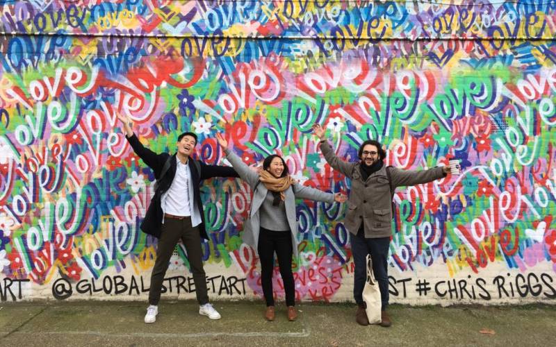 Three students posing in front of graffiti-covered wall (word love written many times in many different colours)