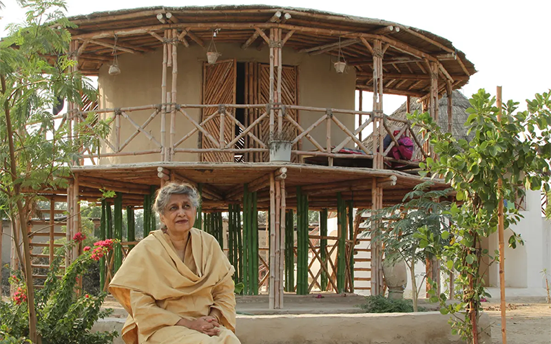 Woman in Pakistani dress sits in front of a circular home on stilts built with traditional materials