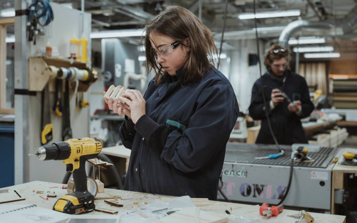 A female student creating a model in the Bartlett workshop, with drills and machinery around her, and a male student working in the background