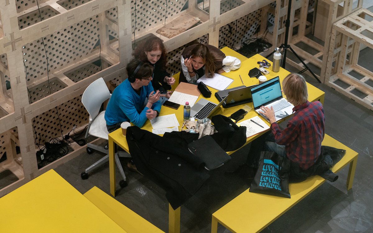 students sit around yellow table with laptops and notebooks, working together on project