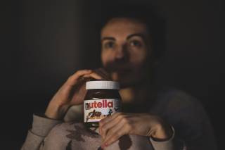 Someone in a low light room with a jar of nutella on their knee