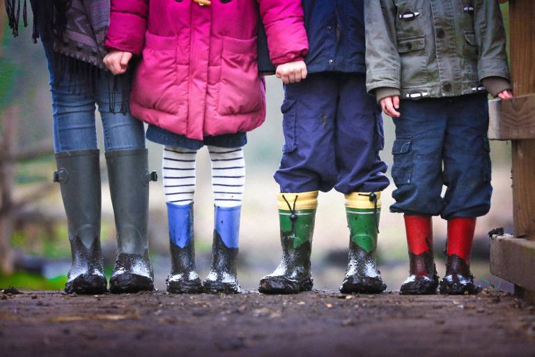 Image is of 4 children, wearing wellington boots. The image is of their lower waist as they stand in a line in front of a puddle.