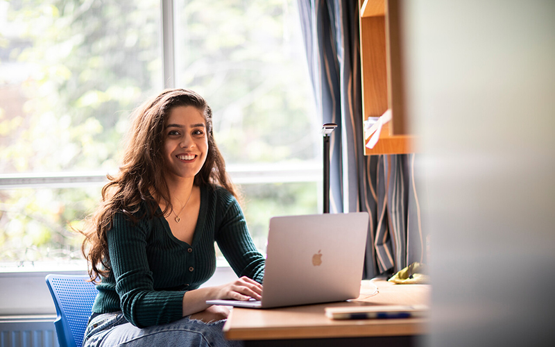A student sat at a desk in University College London accommodation, smiling and in front of a laptop.