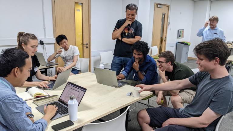 Researchers sit and stand around a laptop on a table.