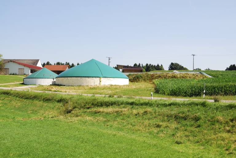 Biogas plant surrounded by green fields and crops, with houses and buildings in the background.