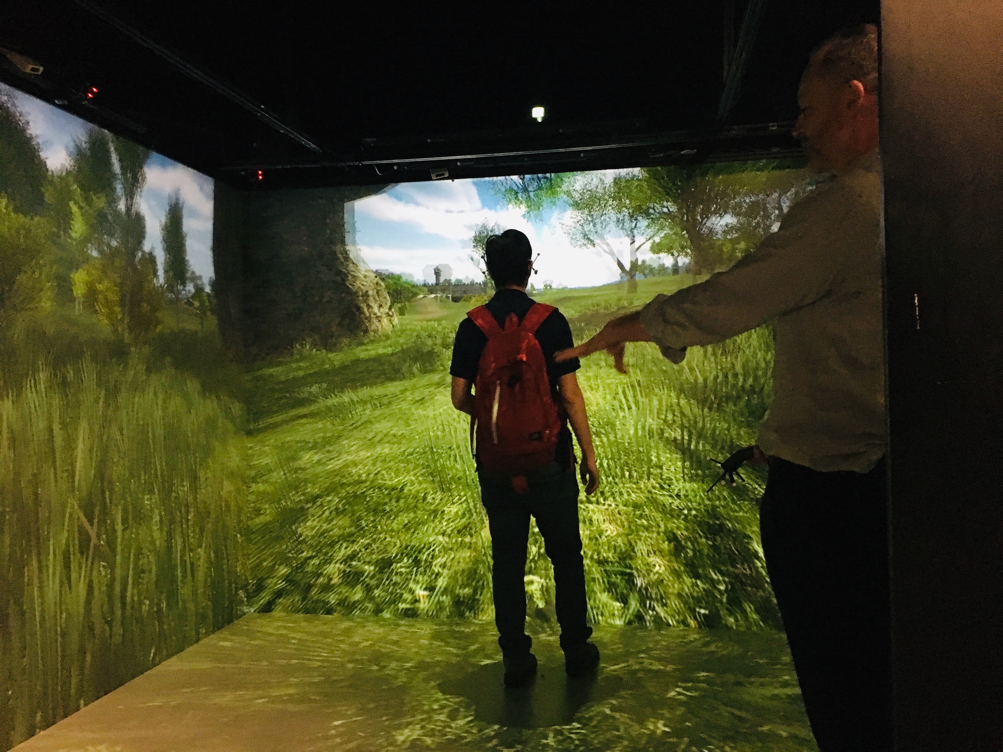 Student wearing backpack using virtual reality cave  displaying an open field landscape with tutor pictured to the side.