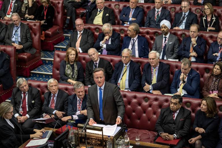 Foreign Secretary David Cameron is stood at the despatch box in the House of Lords. Other members of the Lords are sat next to and behind him, listening to him speak.