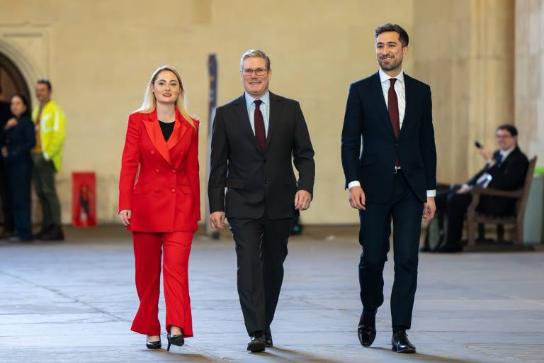 Keir Starmer is walking in a big open indoor space, with a younger, shorter woman walking alongside him on his right, and a younger, taller man walking to his left.
