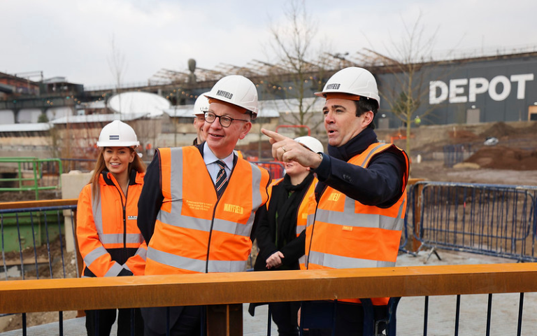 Michael Gove and Andy Burnham in orange high-vis jackets and white hard helmets. Burnham is pointing at something