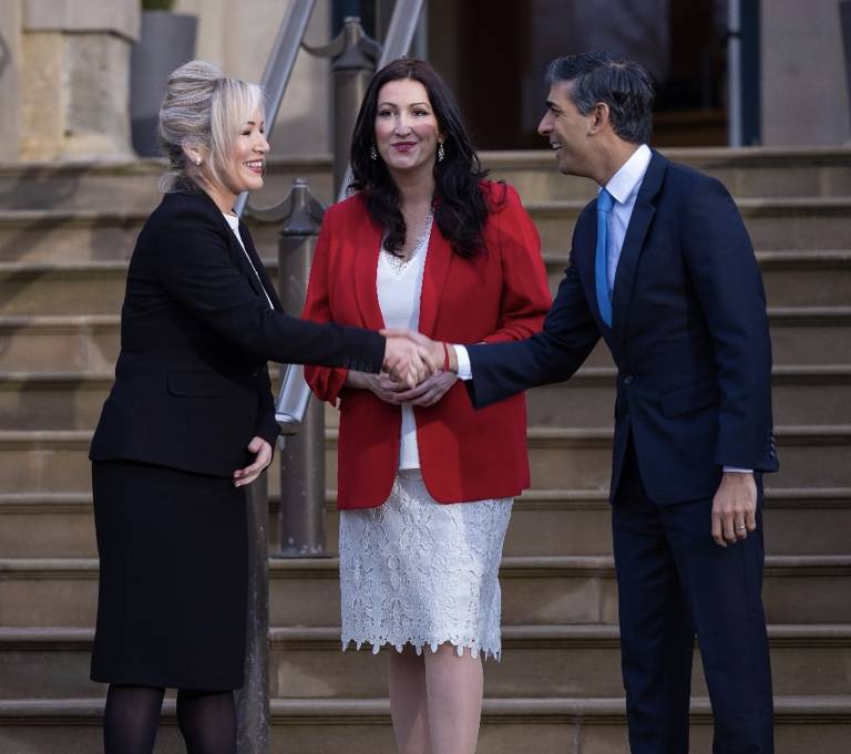 Rishi Sunak shakes hands with First Minister Michelle O’Neill. Both are smiling and side on to the camera. Slightly further back, deputy First Minister Emma Little-Pengelly is stood facing the camera, watching the handshake.