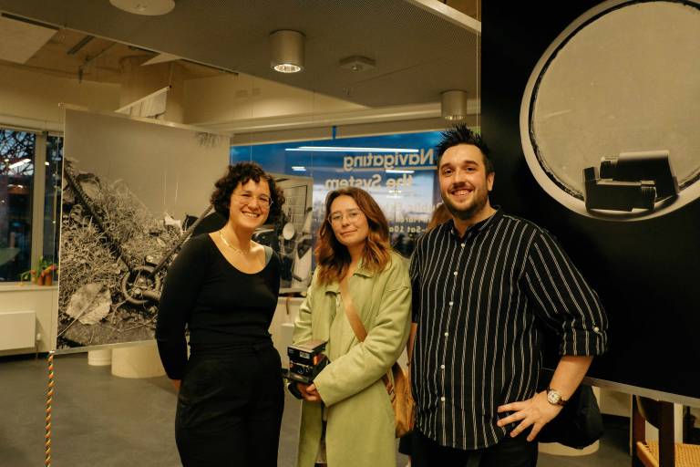 Research participant and boater Caitlin Vinicombe holding a digital camera with UCL researchers Joseph Cook and Nura Ali. smiling and standing in front of the 'Navigating the System' exhibition at the UCL Urban Room in Stratford, East London
