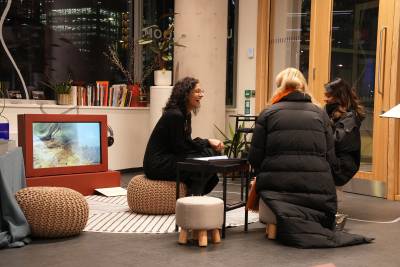 3 people sitting on cushions laughing and talking in exhibition space with books, screens and audio equipment