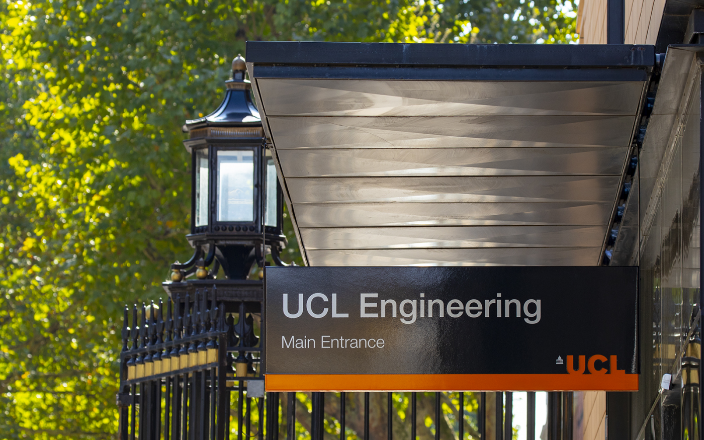 UCL Engineering building main entrance signage. In the background the old classic street lighting, green trees and tall black gated entrance.