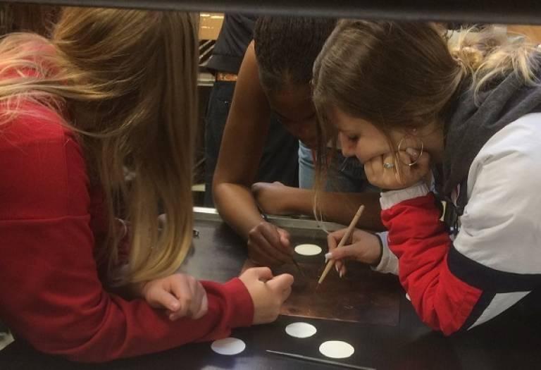 three girls working together on a piece of artwork. Their heads are lowered and elbows resting on the table. They are using tools to make imprints and draw on the material.