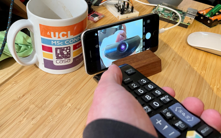 Photograph of a hand pressing a button on a remote control that is pointing towards a phone with the camera function open. On the screen you can see the LED light at the front of the remote and the hand'. 