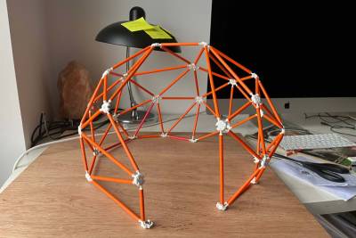 model of geodesic dome made from orange straws and white tape at joints. Model sits on a table with black lamp behind.